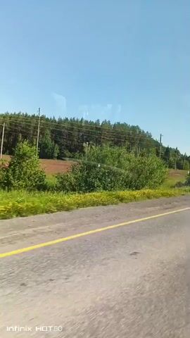 The video is taken from a moving vehicle on a road with a yellow line. The scenery outside includes green grass, trees, and a clear blue sky. There are also power lines and poles visible. A dark-colored car is seen driving in the same direction.