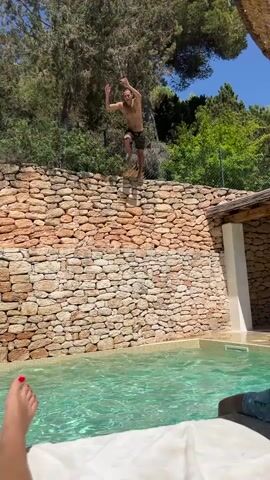 This is a short video clip showing a man jumping into a swimming pool. The man is shirtless and wearing patterned swim trunks. He is in mid-air, with his arms outstretched, just before he enters the water. The pool is surrounded by a stone wall, with lush green trees and vegetation visible behind it. The sun is shining brightly, indicating a warm, clear day. The water in the pool is a vibrant turquoise color. In the foreground, a person's feet are visible, suggesting they are lying down and filming the scene. The overall atmosphere appears to be one of leisure and fun, possibly at a holiday resort or private residence. There is no visible text in the video.