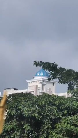 This is a vertical video of a tall apartment building with a blue dome on top. The sky is cloudy and gray. The building is white with balconies on each floor. There are trees in the foreground. The video is shaky and appears to be handheld. There are no people visible in the video.