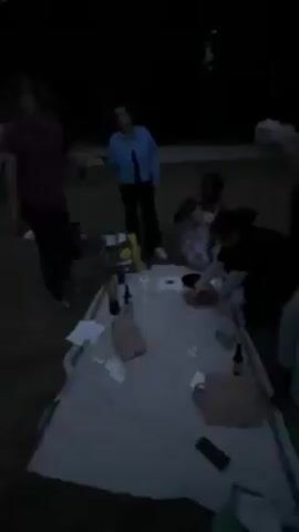 The video begins with a blurry shot of a group of people gathered around a table at night, with various items scattered on the surface. The camera then pans out to reveal a park-like setting under a dark blue sky, possibly dusk or dawn. The scene consists of a grassy area, a paved path, trees lining the horizon, and a building in the distance. The lighting appears dim, with possible lights illuminating the distant building. The video seems to be taken while moving, leading to a slightly shaky view.