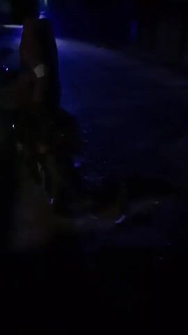 The video shows a dimly lit outdoor area at night. The ground appears to be paved or made of a hard surface. There's a blue light illuminating the scene, which could be from a nearby building or streetlight. The camera moves around, revealing a structure that seems to be a partially constructed or abandoned building with wooden supports. There's no clear indication of people or any specific activity. The setting appears somewhat desolate and possibly an unfinished construction site.