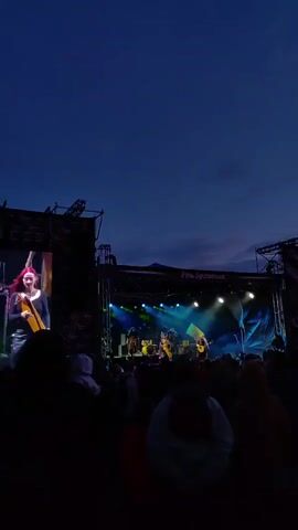 A concert is taking place outdoors at dusk. A large screen on the left displays a close-up of a male performer with green hair wearing a plaid jacket. On the stage in the background, a band is performing under stage lights. A crowd of people are silhouetted in the foreground, facing the stage. The sky is dark blue with a hint of clouds and the setting sun.