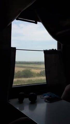 A first-person perspective video shows a view from inside a train car. A table in the foreground holds two dark spherical objects, a blue mask, and a smartphone. Through the open train window, a rural landscape unfolds, featuring fields and trees under a cloudy sky. The video is short and shows no discernible action or interaction.