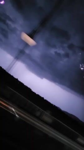 The video displays a dark, blurry, and reflective scene, consistent with a view from inside a moving vehicle at night. The upper portion of the frame is largely black. Throughout the footage, several bright, out-of-focus light sources, appearing as circular white or light-colored blurs, briefly enter and exit the frame, indicative of passing exterior lights. In the lower section, horizontal streaks of light, some orange and some white, are visible, suggesting rapid motion. A faint, blurry pink crescent shape appears briefly in the upper left corner. No discernible individuals or specific objects are clearly visible, only light patterns and reflections. A faint vocalization, "yeah," is audible.