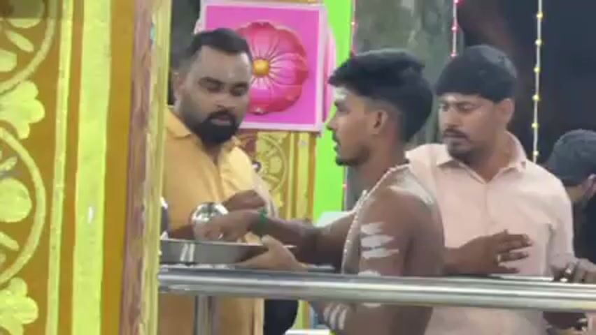 The video shows three men standing inside what appears to be a Hindu temple or shrine. The main subject, a young man with dark hair and white markings on his bare chest and arms, is performing a ritual. He's holding a silver tray with small, reflective objects and a small candle.  He is making gestures with his right hand toward a yellow pillar with painted designs. Two other men are standing behind him. The man directly behind him is wearing a yellow shirt and has a beard. The man on the right has a light pink shirt and is watching the ritual. The backdrop consists of a wall with various colorful decorations and patterns. There is also a fourth person standing on the left who is not clearly visible.