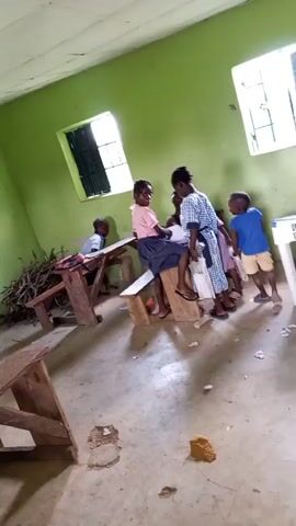The video shows a group of young children in a room with light green walls and two windows with barred openings. The floor is bare concrete. There are wooden desks and benches in the room, some of which appear old and damaged. On the left side of the room, a pile of branches is stacked against the wall. Several children are visible, some sitting on benches and others standing. One child in a pink shirt and blue skirt is sitting on a bench. Another child in a blue and white checkered dress is sitting on the same bench. A child in a blue t-shirt and shorts is standing to the right, looking at the camera. Two other children are in the foreground, one in a black t-shirt and the other in a checkered shirt. There is also a white table with some writing on it in the background. The children appear to be interacting with each other, with some looking like they are playing or talking. The overall impression is that of a simple classroom or gathering space in a basic environment.