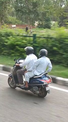Two individuals are riding on a blue scooter. Both are wearing helmets and light-colored clothing. The scooter is in motion on a paved road, with a green fence and trees in the background. The camera appears to be moving alongside the scooter, creating a motion blur effect.