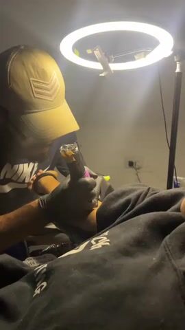 The video shows a close-up of a tattoo artist working on a person's arm. The tattoo artist is wearing a tan baseball cap and black gloves. They are holding a tattoo gun, which is pointed at the person's arm. The person receiving the tattoo is wearing a black hoodie. A ring light is positioned above the tattoo artist, illuminating the workspace. The background appears to be a dimly lit room. The video is very short, showing a brief moment of the tattooing process.