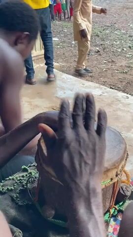 In an outdoor setting in Ogbu, Nigeria, a man is actively playing a traditional drum. He uses both his hands and a stick to strike the drumhead, creating a rhythmic sound. The drum has a natural skin head and appears to be bound with ropes or string. The drummer is shirtless, wearing dark trousers, and is seated on a low surface. Another man is partially visible next to him, seemingly observing.

In the background, a gathering of people, including several children, can be seen standing on a dirt ground beyond a paved area. One man in the background wears a light-colored traditional outfit. A concrete pillar is also visible on the left side of the frame, suggesting a porch or veranda. The scene appears to be taking place during the daytime. No discernible text is visible.