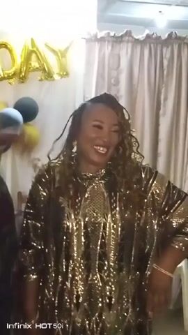 A woman with tan skin and shoulder-length curly hair smiles and poses in front of a backdrop for a birthday celebration. She is wearing a loose-fitting gold sequined top or dress. Behind her, gold balloon letters spell out "DAY" on the wall. To the left of the woman is a bouquet of balloons in black and yellow. The background includes a beige curtain, a wooden chair, and glimpses of other people partially visible on the far left. The video seems to be shot indoors with the logo of "Infinix HOT50i" visible at the bottom left of the screen.
