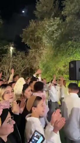 The video shows a crowd of people outdoors at night, likely at a celebration or gathering. Many are raising their arms and clapping, seemingly enjoying the music and the atmosphere. There are trees in the background and a speaker is visible on the right side of the frame. The moon can be seen in the sky. Some of the people are wearing head coverings, and there is a diverse range of ages present.