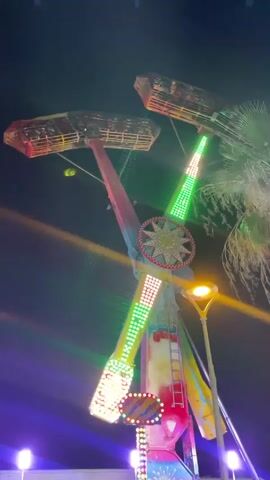 This is a video clip of a carnival ride at night, possibly in Canet de Mar, Spain. The ride is a tall, multi-armed structure with colorful lights. Two gondolas, each holding several people, are attached to the ends of two opposite arms. The ride is in motion, with the arms swinging and rotating. The scene is lit by the ride's lights, streetlights, and the night sky. There is a palm tree visible in the background. The overall mood is festive and exciting. The time of day is clearly night, and the weather appears to be clear. There are no visible words or text in this clip.