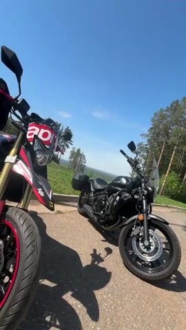 The video shows two motorcycles parked on an asphalt road, possibly a scenic overlook, on a sunny day. One motorcycle, partially visible on the left, is red and white with "Aprilia" written on it. The other is a black cruiser motorcycle with a small windshield and saddlebags. In the background, there is a vast landscape with trees, grass, and hills. The sky is blue with a few clouds. The camera moves slightly, showing the landscape in more detail.