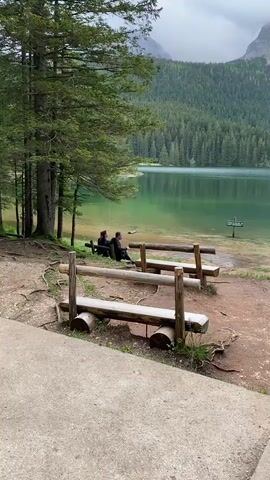 The video captures a serene natural landscape featuring a clear, greenish-blue lake bordered by a dense coniferous forest and surrounded by majestic mountains, some with visible snow patches, under an overcast sky. The setting appears to be a natural park or reserve, with rustic wooden benches by the water.

In the foreground, a concrete pathway leads to a reddish-brown dirt path where two wooden benches, made from logs, are placed. On the left bench, two men are seated, facing the lake and observing the scenery. To the right, three women stand near the water's edge, also looking out at the lake or conversing. A dark green sign with illegible text and a blue circular sign with a white symbol are visible in the shallow water near the women.

The lake's water is clear enough to show the lakebed near the shore and reflects the surrounding trees and sky. Large pine trees frame the left side of the shot. The dominant colors are the vibrant green of the forest and lake, the gray of the sky and mountains, and the reddish-brown of the earth. The overall activity involves people relaxing, observing the beautiful natural environment of the lake and mountains during what appears to be a cloudy daytime. No clear text is legible on any of the signs.
