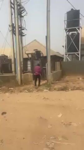 A man in a pink shirt and dark pants is standing next to an electrical transformer and a concrete utility pole. He is wearing a safety harness. The transformer is mounted on a metal frame behind a metal gate in front of a building with a corrugated metal roof. Behind the transformer is a large water tank. The ground is dry and dusty. The sky is hazy.