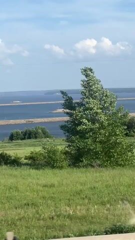 The video shows a view of a landscape from a moving vehicle, likely a car. The foreground features a grassy field with green trees swaying in the wind. In the background, there's a wide body of water with what appears to be a bridge or land connecting different parts of the water. The sky is blue with fluffy white clouds. An electrical power pole briefly appears in the middle of the frame.