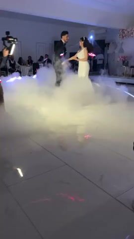 In a well-lit banquet hall, a newlywed couple is engaged in their first dance, surrounded by an abundance of fog. The room is decorated with white marble flooring and illuminated by LED lights that project vivid pink, blue, and purple hues, enhancing the romantic atmosphere. The groom is dressed in a classic black suit, and the bride is radiant in a white wedding gown, with a tiara accentuating her elegant style. As the music plays, the groom gently dips his bride, symbolizing their unity.

Several guests are seated at tables along the edges of the dance floor, observing the couple and participating in the joyful celebration. Background elements of the scene include a DJ booth and additional event setup, indicating a lively party atmosphere.