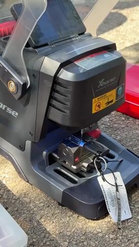 The video shows a close-up of a black and gray Xhorse automatic key cutting machine outdoors. The machine has a clear protective shield over the cutting area. A key is clamped in the machine, and a cutting tool is actively working on it, creating sparks. A tag attached to the key reads "PIN CODE PROCEDURE" and includes a barcode and serial number. The background is out of focus, but appears to be a paved surface, possibly a driveway or sidewalk, with some greenery visible. A red case is partially visible on the right side of the frame. The lighting suggests it is daytime, and the presence of shadows indicates sunlight. The machine is positioned on the ground. There is no visible text on the machine itself beyond the brand name "Xhorse" and a warning sticker. The overall impression is of a functional, in-progress key duplication or programming task.