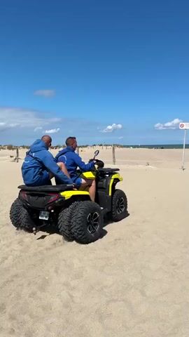 Two men ride on a yellow ATV on a sandy beach. They are both wearing blue hooded sweatshirts. The ATV is moving away from the camera, kicking up sand. In the background, the ocean is visible under a blue sky with some clouds. There are a few people and beach umbrellas further down the beach. A sign is visible on a pole to the right of the ATV.