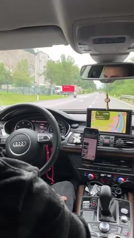 The video provides a first-person view from the driver's seat of a car. The driver's hands are visible on the steering wheel. The dashboard displays a speed of 78 km/h. A phone is mounted on the dashboard and appears to be playing a video. The car's navigation system is also displayed, showing a route with 3.8 km remaining. Outside the car, the scenery is green with trees and a road. A truck and a motorcycle are visible on the road ahead. A rearview mirror shows the reflection of the driver's face. The interior of the car appears to be well-maintained with various accessories, including a rosary hanging from the rearview mirror and small teddy bear decorations on the dashboard. The weather appears to be overcast.