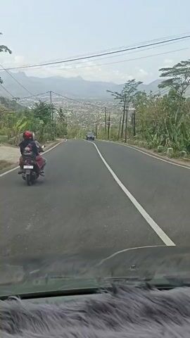 The video shows a downward sloping paved road with a white line dividing the lanes. The perspective is from inside a vehicle looking forward. On the left side of the road, a motorcycle with two people riding on it is visible, moving away from the camera. Further ahead on the road, a dark-colored car is moving in the same direction. On the right side of the road, there are trees and utility poles. In the distance, a town or city is spread out below, with mountains in the background. The sky is clear and bright.