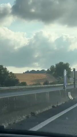The video shows a first-person perspective of someone driving on a highway. The road is paved with asphalt and has white lane markings. There is a concrete barrier on the left side of the road. In the distance, there are rolling hills covered in vegetation and fields. The sky is cloudy with patches of blue visible. There are a few cars driving on the highway ahead. There are also several road signs visible. One sign on the left appears to be a speed limit sign. Another sign on the right indicates a distance to a destination. The overall scene suggests a journey through a rural or semi-rural area.
