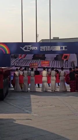 A group of six women dressed in white gowns with their backs to the camera are standing in a line, holding a red banner. They are facing a large screen that displays an image of what appears to be a vehicle. Behind the women, several men are standing, some of whom are also holding the banner. To the right of the group, a person in a red and black costume, possibly a lion dance costume, is visible. The background features a large blue banner with the word "CARRY" and Chinese characters printed on it. The scene appears to be outdoors, with a paved ground and what looks like a building or structure in the background under a bright sky. The overall setting suggests an event or a ceremony, possibly a product launch or a company event.