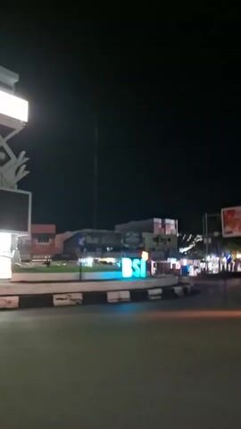The video shows a night time view of a roundabout with a large, illuminated monument in the center. The monument has a square top section with bright white lights and a large, dark screen below it. The base of the monument is decorated with white, abstract shapes that resemble leaves or petals. In front of the monument, large, blue, illuminated letters spell out "BSI". The roundabout itself is marked with black and white lines. In the background, there are buildings and streetlights, indicating an urban environment. The sky is dark, suggesting it is nighttime. The camera appears to be moving, possibly from a vehicle, as the foreground is blurred.