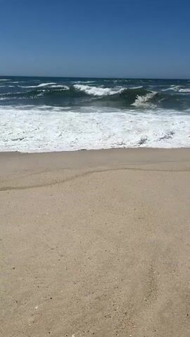 This is a vertical video of a beach scene in Torreira, Portugal. The video captures a clear, blue sky with no clouds. The ocean is visible with moderately sized waves breaking and creating white foam that washes onto the sandy shore. The sand appears light brown and has some subtle patterns, possibly from the receding tide or wind.

The scene is set during the daytime, likely mid-morning to early afternoon, given the bright sunlight and clear sky. The weather appears to be warm and sunny, typical for a coastal summer day. The gentle crashing of the waves and the vast expanse of the ocean suggest a peaceful and serene atmosphere, evoking a sense of relaxation and the natural beauty of the seaside.

There are no people or man-made objects prominently featured in the video. The focus is purely on the natural elements: the sky, the sea, and the sand. The video is a snapshot of a tranquil moment at the beach, highlighting the dynamic interaction between the waves and the shore. There is no visible text in the media file.