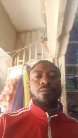 The video shows a man taking a selfie in what appears to be a small store. The man has a beard and is wearing a red jacket with white stripes on the shoulders and a cap. Behind him, shelves are stocked with various products, including food items, and on the left, there is an advertisement poster. Two other men are visible in the background. The video is slightly shaky.