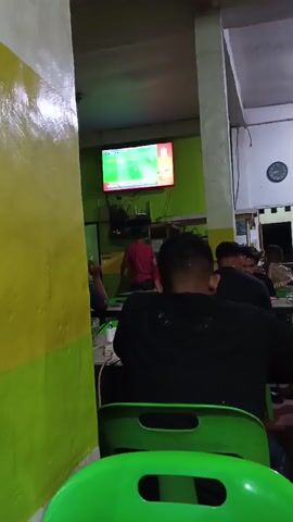 The video shows a group of people sitting at tables in what appears to be an indoor eating establishment. A television screen is visible on the wall in the background, displaying a sports broadcast. The lighting is dim, and the walls are painted green and yellow. Several individuals are seated, facing away from the camera, some are wearing black shirts. One person is standing and gesturing towards the front of the room. The overall atmosphere seems casual, with people engaged in watching the television or conversing.