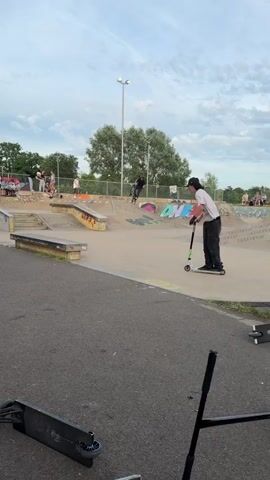 Here's a description of the video:

The video shows a person performing a trick on a scooter at a skatepark. The individual, wearing a white shirt, black pants, and a black baseball cap, rides the scooter towards a concrete ledge. They jump, landing with both feet on the ledge and then hopping back down onto the ground. 

The skatepark has various ramps and concrete structures, with graffiti visible on some surfaces. Other people are present in the background, some watching the scooter rider. A black scooter lies on the ground in the foreground. The sky is overcast.