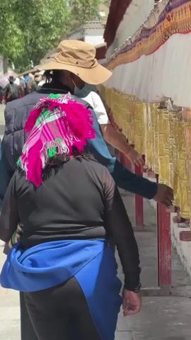 A group of people walks outdoors alongside a long white wall lined with numerous golden prayer wheels. The wall has a red base and colorful decorative banners or fabrics running above the prayer wheels. The person in the foreground, seen from behind, wears black clothing, a bright pink and green patterned headscarf, and a blue fanny pack or draped fabric around their waist. Ahead of them is another person wearing a brown hat and a dark vest. Further down the path, a person in a white top and a red baseball cap is visible. Most of the individuals are using their right hands to spin the prayer wheels as they pass, engaging in a common Buddhist ritual. The scene is brightly lit by sunlight, indicating daytime. Trees and more distant buildings can be seen in the background.