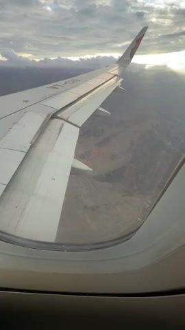 The video shows a view from an airplane window looking down at a barren, mountainous landscape. The airplane's wing is visible in the foreground, and a long, winding road or riverbed stretches across the desert terrain below. The sky is cloudy with sunlight breaking through.