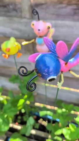 This is a close-up video of three decorative garden stakes in a garden setting. The stakes are shaped like whimsical insects and a bird. In the foreground, a blue and pink metallic dragonfly-like figure is on a green stake. It has large black eyes with white highlights, black curly antennae, and iridescent blue wings with pink tips. Behind it, slightly out of focus, is a pink metallic bee-like figure with yellow accents and light blue detailing on its body, also on a green stake. It has yellow wings and appears to be waving. To the left, a bright yellow metallic bird with orange feet and beak is perched on a green stake. All three figures are positioned amongst lush green plants and foliage, suggesting a garden environment. The background consists of a weathered wooden fence, indicating an outdoor setting. The lighting suggests it is daytime. The overall mood is playful and cheerful, conveyed by the colorful and animated designs of the garden ornaments. There is no visible text in the video.