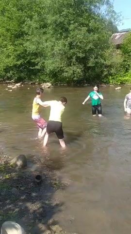 Three young people are playing in a shallow river. One person, wearing a green t-shirt and black shorts, stands in the water. Another person, wearing a yellow t-shirt and black shorts, appears to be wrestling with a third person, who is wearing a purple and pink outfit. The river has a muddy bottom and is surrounded by lush green trees and vegetation. The sky is clear and sunny.