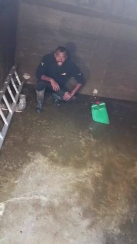 A man is crouching in a flooded, square, concrete enclosure. He's wearing a black Puma sweatshirt and black rubber boots. Next to him is a green jerrycan with a red cap. A ladder is propped against the wall to his left. The floor is wet and covered in a layer of water and grime. The lighting is dim. The man is looking towards the camera. Distant chanting sounds are audible.