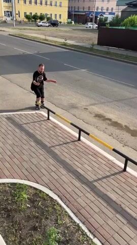 A person is rollerblading on a metal railing alongside a sidewalk and a street. The person is wearing a black t-shirt and black shorts. There is another person standing on the sidewalk holding a camera, filming. The scene takes place outdoors during the daytime, with buildings and trees visible in the background.