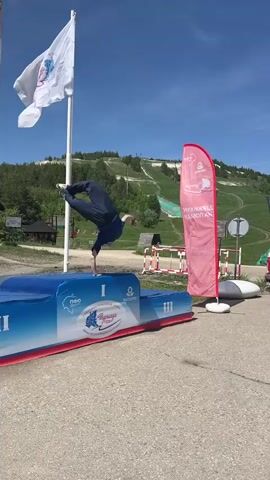 A man in a blue shirt and dark pants is performing a parkour move in an outdoor setting with a ski slope visible in the background. He vaults over a long, blue and red structure with white lettering on it. To the left is a flag with a white background and a circular emblem. To the right is a red banner with white text and a white emblem, and behind that is a red and white VW bus. The man lands on his feet and takes a few steps. The sky is clear and blue. There are some trees on the side of the ski slope. The ground is paved.