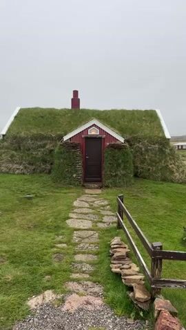 A traditional Icelandic turf house is centered in the frame, set against an overcast sky. The small dwelling features red-painted wooden walls, a dark brown door, and a prominent, thick green turf roof from which a red chimney rises. White trim outlines the eaves of the roof. The turf extends down the sides of the house, blending the structure with the surrounding green grass.

A rustic pathway of irregular flat stone slabs leads from the foreground directly to the front door. To the right of the path, a low, dark wooden fence runs partially into the frame, with a small pile of natural stones gathered near its base. The ground in the immediate foreground is a mix of gravel and green grass.

The scene depicts a tranquil, rural setting, characteristic of the architecture found in Bakkagerði, Iceland. The cloudy, muted light suggests an overcast day. No people are visible, and there is no discernible text in the image.