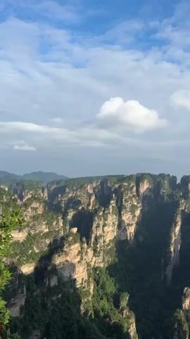 The video shows a scenic landscape of Zhangjiajie National Forest Park in China. Tall, slender sandstone pillars covered in green vegetation rise from a deep valley floor. The sky above is partly cloudy, with patches of blue visible. The camera slowly pans across the vista, revealing the vastness and unique geological formations of the area.