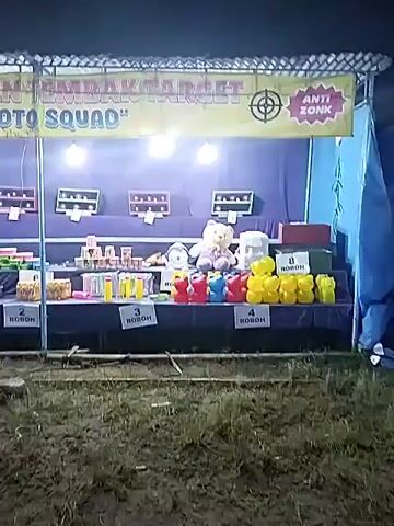 The video shows an outdoor game booth at night, with several people standing around. The booth is decorated with a sign that reads "IN TEMBAK TARGET 'OTO SQUAD' ANTI ZONK". There are various prizes displayed, including stuffed animals, boxes, and small toys, arranged on a table. The scene is lit by bright overhead lights. People are visible on a wooden platform in front of the booth, some are standing and others are sitting. The ground in front of the booth is uneven and covered with grass. The camera movement is somewhat shaky.