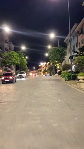 A first-person perspective video shows a street at night. The street is paved with interlocking rectangular stones. On the left side of the street are trees and a white car. On the right side of the street are buildings with lights on. There are streetlights that cast a glow on the street. A red vehicle with multiple windows, resembling a trolley, drives down the street from the left side toward the center. Other cars are visible in the distance, also driving on the street. The buildings are lit up with lights and have balconies and trees in front of them. The overall scene is one of a bustling street at night, possibly a commercial or tourist area.