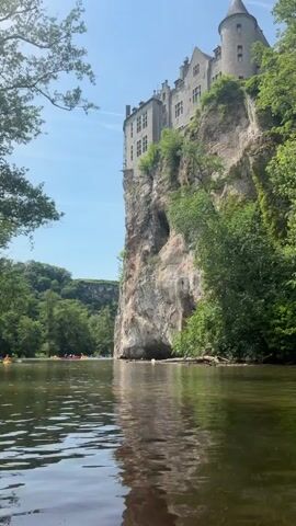 The video shows a castle perched atop a cliff overlooking a body of water. The castle is a multi-story stone structure with a turret and several windows. The cliff is rocky and covered in vegetation, and there are trees on either side of the castle. In the water, there are several kayaks with people paddling in them. The sky is blue and there are a few clouds visible. The water is calm and reflects the sunlight.