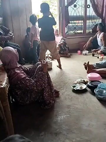 A group of people are gathered in what appears to be a home. In the foreground, a woman wearing a headscarf and a patterned dress is sitting on the floor, facing away from the camera. To her left, partially visible, is another person also seated. In the center of the room, a young boy in a dark shirt and light-colored shorts is standing and appears to be interacting with something on the floor. Behind him, another child in a pink shirt is also standing. To the right, several other individuals are seated on the floor. One man in a white tank top is visible. There are various objects scattered around the floor, including plates with food, cups, and other items. The room has wooden walls and a large window with curtains. Outside the window, trees and a vehicle are visible. The lighting suggests it is daytime. The overall atmosphere seems informal, possibly a family gathering or a social occasion.