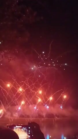 A fireworks display is taking place over a body of water at night. Numerous red and orange fireworks are exploding in the sky, creating trails of light and smoke. In the foreground, the silhouettes of several people can be seen watching the display. One person is holding up a smartphone to record the event. The water reflects the lights of the fireworks.