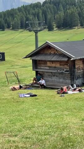 A group of people are relaxing on a grassy hillside next to a wooden building. There are at least five individuals visible. One person is lying on their back on a blanket, another is lying on their stomach on a towel, and a third is lying on their side on a blanket. Two people are sitting on steps leading up to the wooden building. The building appears to be a small, rustic structure with a dark roof and wooden walls. In the background, there is a ski lift tower and a dense forest. The landscape is mountainous with a clear sky and scattered clouds. A black soccer-style net frame is set up on the grass to the left of the building.