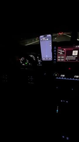 The video shows the interior of a car at night, focusing on the dashboard and a smartphone mounted on the windshield displaying a navigation app. The driver's side is visible, with the steering wheel and instrument cluster illuminated. The speedometer shows a speed of 132 km/h. The central infotainment screen is also visible, displaying music information. The car is traveling on a highway, as indicated by the illuminated lane markers and the distant headlights of other vehicles. The navigation app on the phone shows a route with 130 km remaining. The overall environment is dark, with the interior lights of the car providing the primary illumination.