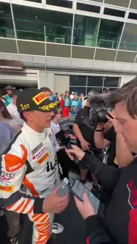 A male race car driver in a white and orange racing suit with "PIRELLI," "TRESOR," and "Audi Sport" logos is being interviewed outdoors at what appears to be a racetrack in Monza, Italy. He wears a black Pirelli cap with "1ST" embroidered on it and is holding a small water bottle. He is adjusting his cap while looking towards the interviewer. The interviewer, dressed in black, holds a microphone with a red logo close to the driver, and a cameraman is filming the interaction with a professional camera and a large lens. In the background, a crowd of spectators is visible, along with a modern building featuring large windows and a sign that partially reads "NGINEERING GT2/GT4". The scene is bright and sunny, suggesting daytime. The driver appears calm and engaged in the interview.