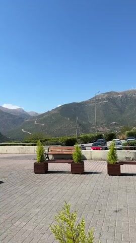 The video shows a winding road up a green, mountainous landscape under a clear blue sky. In the foreground, there is a paved area with planters containing small evergreen trees and a wooden bench. A white barrier separates the paved area from the natural landscape. Cars are visible driving on a road in the mid-ground.